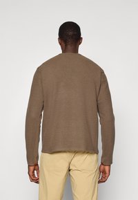 Brauner strukturierter Pullover mit langen Ärmeln und rundem Halsausschnitt. Trägt man über hellbeigen Hosen, die eine lockere Passform und ein Zickzackmuster zeigen.