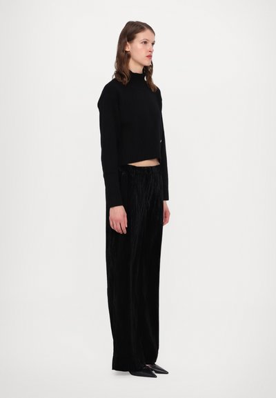 Schwarzer, kurz geschnittener Pullover mit hohem Kragen und geripptem Saum, kombiniert mit schwarzen, samtigenweiten Hosen, die eine glatte Textur und einen subtilen Glanz aufweisen.