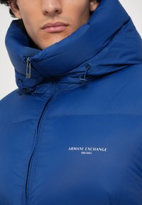 Plava zimska jakna s visokim ovratnikom, sa zatvaranjem na zatvarač i detaljima s vezicom. Na njoj se nalazi logo "Armani Exchange Milano" u bijeloj boji.