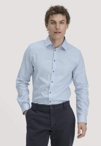 Lichtblauw overhemd met puntige kraag, bruine knopen en een gestructureerde stof. Draag het met donkerblauwe broek.