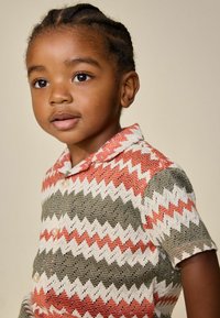 Jedes kleines Kind mit geflochtenen Haaren trägt ein kurzärmeliges Shirt mit Zickzackmuster in Rot, Weiß und Olivgrün und blickt leicht zur Seite.