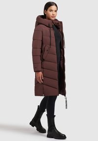 Dlouhý hnědý puffer kabát s velkou kapucí, zipem, dvěma postranními kapsami a diagonálním prošíváním. Nesený přes černé legíny a boty.