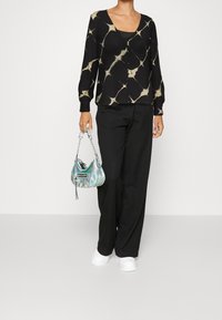 Blusa nera con motivo a disegni astratti beige, abbinata a pantaloni neri larghi e borsa scintillante color teal, insieme a sneakers bianche.