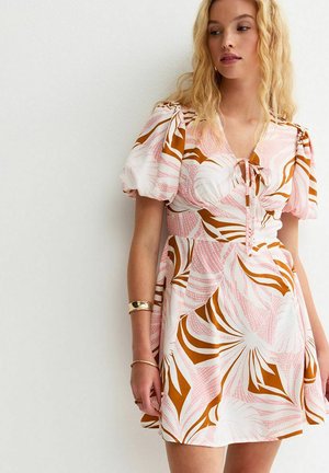 Kobieta w krótkiej, wzorzystej sukience z bufkami na rękawach w kolorach białym, różowym i brązowym, stojąca na tle gładkiej jasnej ściany.