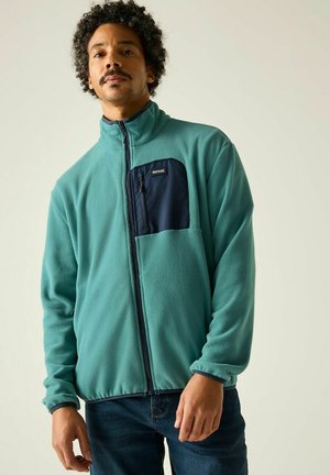 Mann trägt eine türkisfarbene Fleecejacke mit schwarzer Brusttasche und steht vor einem schlichten hellen Hintergrund.