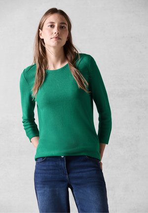 Pull en maille texturée vert avec un col rond et des manches trois-quarts, associé à un jean bleu foncé. Le matériau semble doux et respirant.