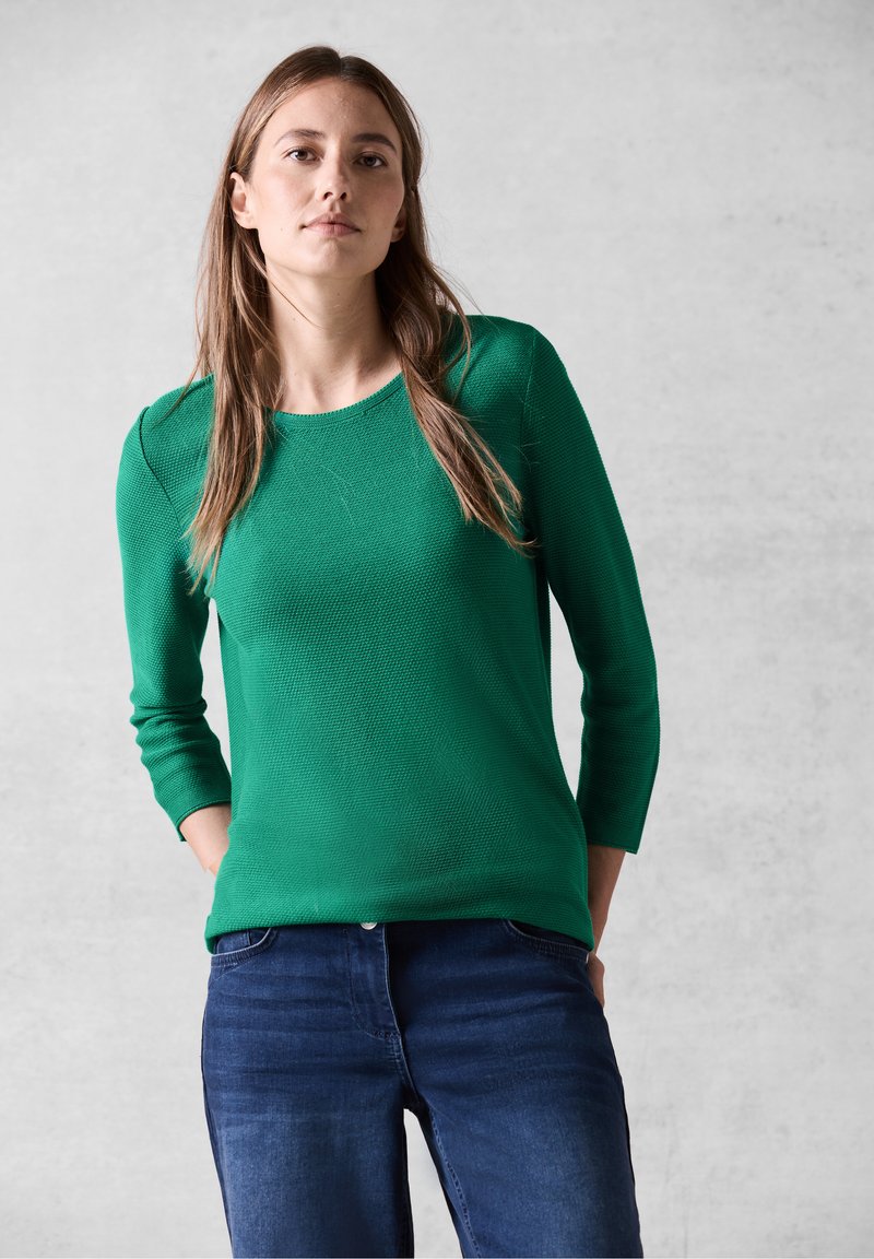 Maglioncino verde a maglia con scollo rotondo e maniche tre quarti, abbinato a jeans blu scuro. Il materiale sembra morbido e traspirante.