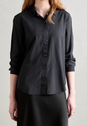 Kobieta ubrana w czarną koszulę na guziki z długim rękawem, ozdobioną subtelnym geometrycznym wzorem, oraz czarną spódnicę na jednolitym tle.