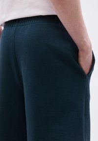 Άτομο που φοράει σορτς με ελαστική μέση σε σκούρο μπλε χρώμα, με το χέρι εν μέρει στην πλαϊνή τσέπη, φοράει ανοιχτόχρωμο πάνω μέρος ενδυμασίας σε απλό φόντο.