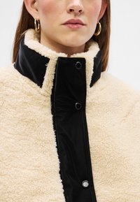 Κρέμ φλις μπουφάν με μαύρο νάιλον μπροστινό πάνελ, που διαθέτει ψηλό γιακά και κουμπιά με κλιπ κατά μήκος της άκρης. Μαλακό, υφασματισμένο ύφασμα.