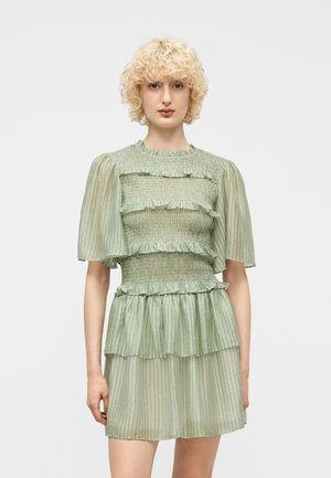 Person med kort, krøllet blondt hår som har på seg en lys grønn, plisssert kjole med ruffled lag og puffede halv-ermer, stående foran en ensfarget bakgrunn.