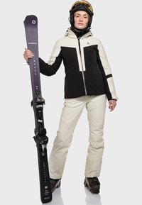 Skiausrüstung mit einer zweifarbigen Jacke in Schwarz und Creme mit Kapuze, cremefarbenen Hosen und schwarzen Skischuhen, die schwarze Blizzard-Ski hält.