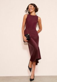 Bordó ujjatlan overall derékban megkötővel, légies sziluettel és oldalsó hasítékkal. Fekete kis táskával és hegyes orrú magassarkúval párosítva.