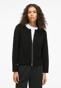 Črna zip-up jakna iz teksturirane tkanine z okroglim izrezom in dolgimi rokavi, v kombinaciji z črnimi in belimi črtastimi hlačami.