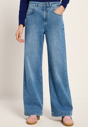 Person trägt hellblaue, weit geschnittene Jeans mit Händen in den Taschen und rosa Sneakers vor einfarbigem Hintergrund.