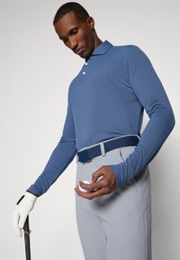 Polo majica s dugim rukavima u plavoj boji s ovratnikom, svijetlosive hlače i tamnoplava kaiša, drži lopticu za golf i palicu, nosi bijelu rukavicu.