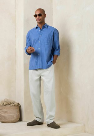 Mand iført blå langærmet skjorte, hvide bukser, solbriller, stående indendørs nær en beige væg med en flettet kurv på gulvet.