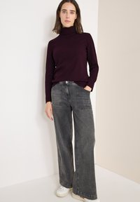 Ciemnofioletowy sweter z golfem w połączeniu z szarymi spodniami z szerokimi nogawkami, posiadającymi naszywane kieszenie i luźny krój, noszony z białymi sneakersami.