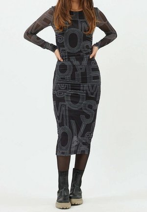 Kobieta w czarnej midi sukience z długim rękawem, ozdobionej dużymi białymi konturami liter, oraz w czarnych botkach z beżowymi podeszwami.