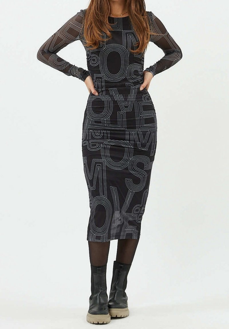 Femme portant une robe midi noire à manches longues avec de grandes lettres blanches en contour et des bottes noires à semelles beige.