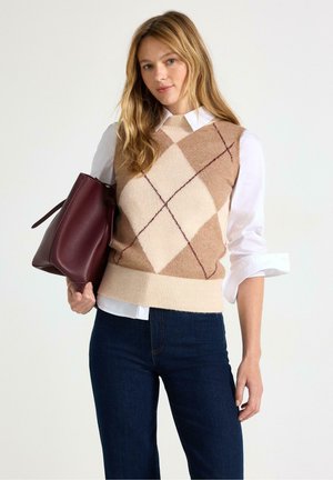 Kvinde iført beige argyle sweatervest over hvid skjorte, mørke jeans, holder en stor mørkerød håndtaske mod en ensfarvet baggrund.