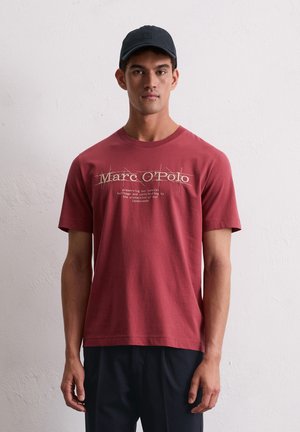 Rotes Baumwoll-T-Shirt mit einem grafischen Druck in Weiß, kurzen Ärmeln und Rundhalsausschnitt. Getragen mit einer dunklen Hose und einer schwarzen Mütze.