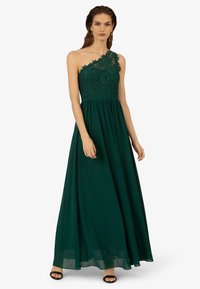 Ein schulterfreies Kleid in einem tiefen Grünton mit Spitzenverzierungen und einer taillierten Passform. Der fließende Rock verleiht Volumen. Das Model trägt schwarze Absätze, die Haare offen frisiert.