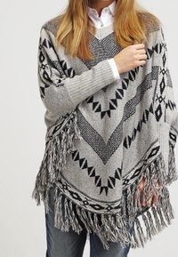 Kobieta w szarym poncho z czarnymi geometrycznymi wzorami i frędzlami, nosząca białą koszulę i niebieskie dżinsy, na jednolitym tle.