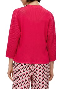 Rotes geripptes Strickoberteil mit Dreiviertelärmeln und Rundhalsausschnitt. Kombiniert mit gemusterten rosa und schwarzen Hosen.