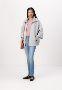 Grå hettejakke med brede ermer, lagd over en rosa stripete skjorte. Lyseblå jeans og svarte leppesko med stropper. Brun crossover-veske.