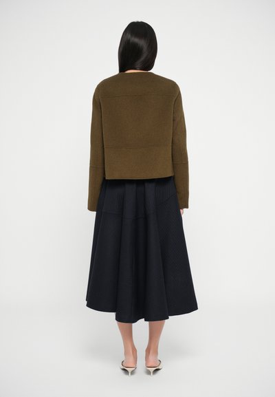 Lühendatud oliivroheline jakk, millel on struktureeritud õlad, koos voolava musta volditud seelikuga, kantud neutraalsete kingadega.