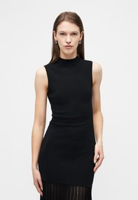 Γυναίκα που φοράει εφαρμοστό μαύρο φόρεμα χωρίς μανίκια με ψηλή λαιμόκοψη και διαφανές διακοσμητικό κρόσι στο τελείωμα, στέκεται μπροστά σε απλό λευκό φόντο.