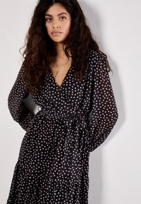Schwarzes Wickelkleid mit weißen Punkten, langen Ärmeln und einer Bindung am Taillengürtel; verfügt über einen gestuften Rock und einen tiefen V-Ausschnitt.
