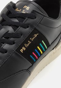 Zapatilla de cuero negra con cordones negros, que presenta un acento a rayas multicolores y el logotipo dorado "PS Paul Smith" en el lateral.