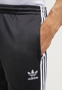 Svarta träningsbyxor med vita ränder på sidorna, med en mjuk textur och en broderad Adidas-logotyp på vänster lår.