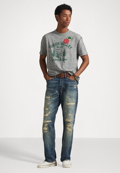 Harmaa T-paita, jossa on vihreä graafinen teksti ja kuvio; siniset kuluneet farkut, joissa on repeämiä; valkoiset kengät; ruskea vyö, jossa on näkyvä solki.