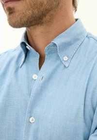 Camicia azzurra chiaro con colletto, dotata di bottoni bianchi e tessuto testurizzato. La vista ravvicinata mette in evidenza i dettagli delle cuciture e del colletto.