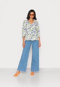 Blusa com padrão floral, com decote em V e mangas 3/4, combinada com calças de ganga de perna larga de cintura alta na cor azul e sandálias de salto bloco laranja.