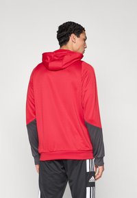 Červená sportovní mikina s šedým akcentem na rukávech, s kapucí na stažení a hladkou texturou. Nošena s černými kalhotami se pruhy.