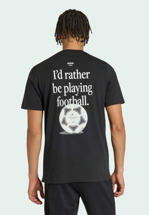 Person mit geflochtenem Haar trägt ein schwarzes Adidas-T-Shirt mit dem Text „Ich würde lieber Fußball spielen“ und einer Fußball-Grafik auf dem Rücken.