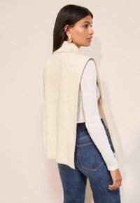 Kreemi lõhthihaga vest pehme tekstuuriga, mille all on pikakäistega valge topp, kombineeritud tume siniste teksadega.