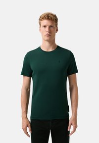 Młody mężczyzna z krótkimi, kręconymi blond włosami, ubrany w ciemnozielony T-shirt z krótkim rękawem i czarne spodnie, stojący na tle białym.