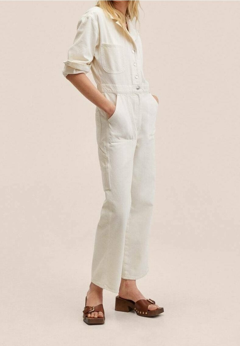 White denim jumpsuit with long sleeves, chest pockets, and button closure, paired with brown platform sandals, set against a neutral background.