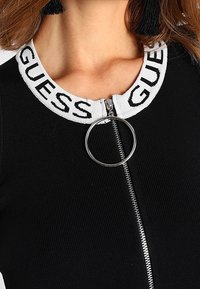 Haut noir zippé avec col côtelé blanc portant le texte "GUESS" et un tirette en anneau argenté sur la fermeture éclair, tissu texturé avec des lignes verticales.