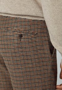 Béžový svetr zastrčený do hnědých, námořnických a krémových vlněných kalhot s kostkovaným vzorem houndstooth a knoflíkovou zadní kapsou.