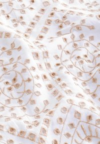 Biały materiał z misternym beżowym haftem, przedstawiającym wzory florystyczne i geometryczne, ukazujący teksturowaną powierzchnię z elementami trójwymiarowymi.