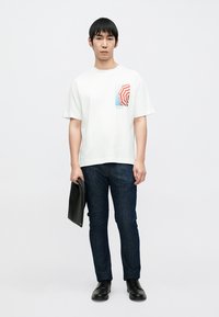 T-shirt blanc en coton avec graphique d'un parapluie rouge et bleu sur la poitrine gauche, associé à un jean bleu foncé et des chaussures noires. Porte un dossier noir à la main.