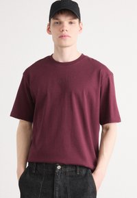 Burgunderfarbendes kurzärmeliges T-Shirt aus Baumwolle, mit einem runden Ausschnitt und gesticktem "BOSS"-Logo auf der Brust. Kombiniert mit schwarzen Denim-Hosen.
