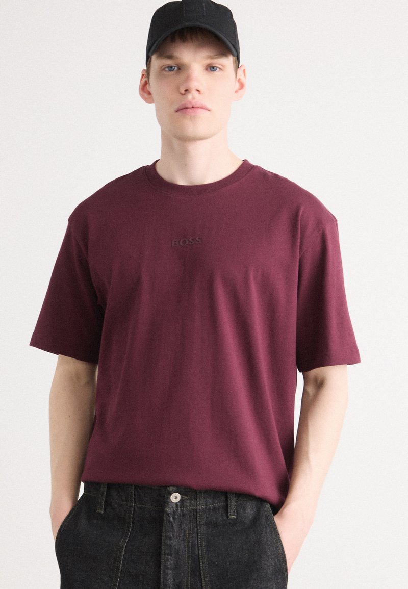 Burgunderfarbendes kurzärmeliges T-Shirt aus Baumwolle, mit einem runden Ausschnitt und gesticktem "BOSS"-Logo auf der Brust. Kombiniert mit schwarzen Denim-Hosen.