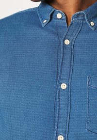 Blå skjorta med fiskbensmönster och knäppning vid kragen, med vita knappar och en bröstficka. Den strukturerade tyget ger djup.